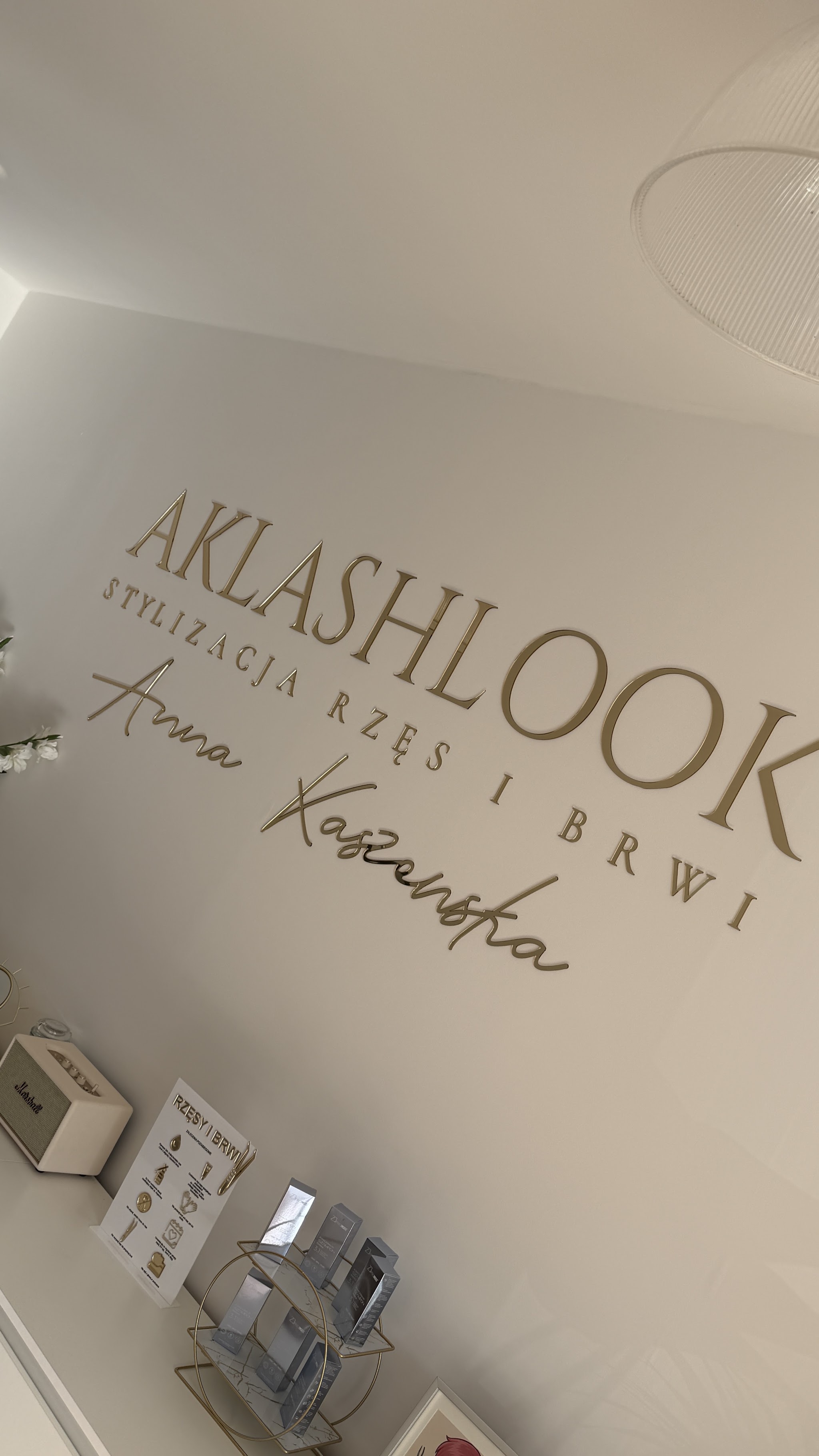 De muur in de salon met gouden letters AKLASHLOOK, Stylizacja rzęs i brwi, Anna Kaszewska, en nazorgproducten op een plank